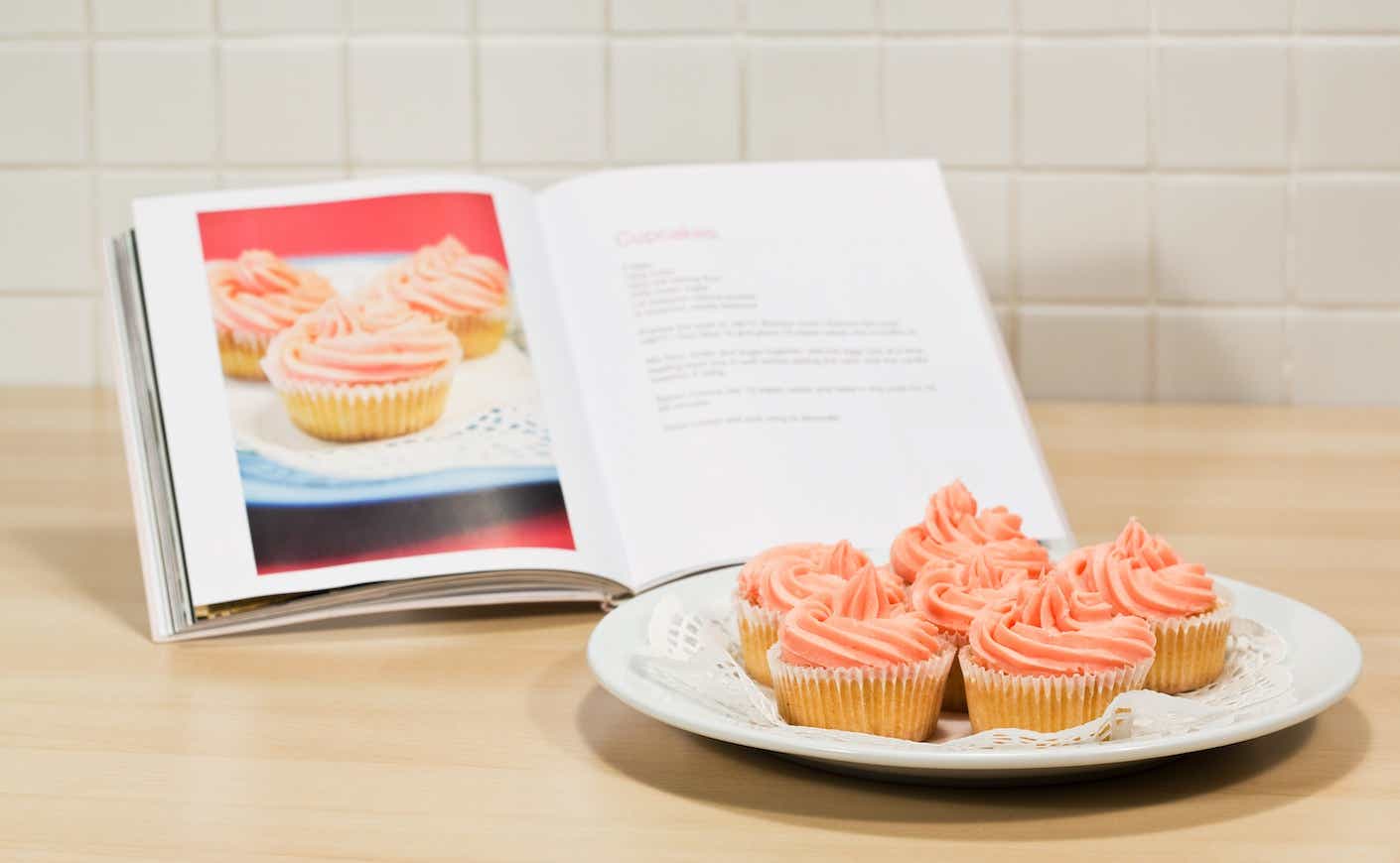 plate of cupcakes with pink frosting in front of a cookbook with a picture of the same cupcakes