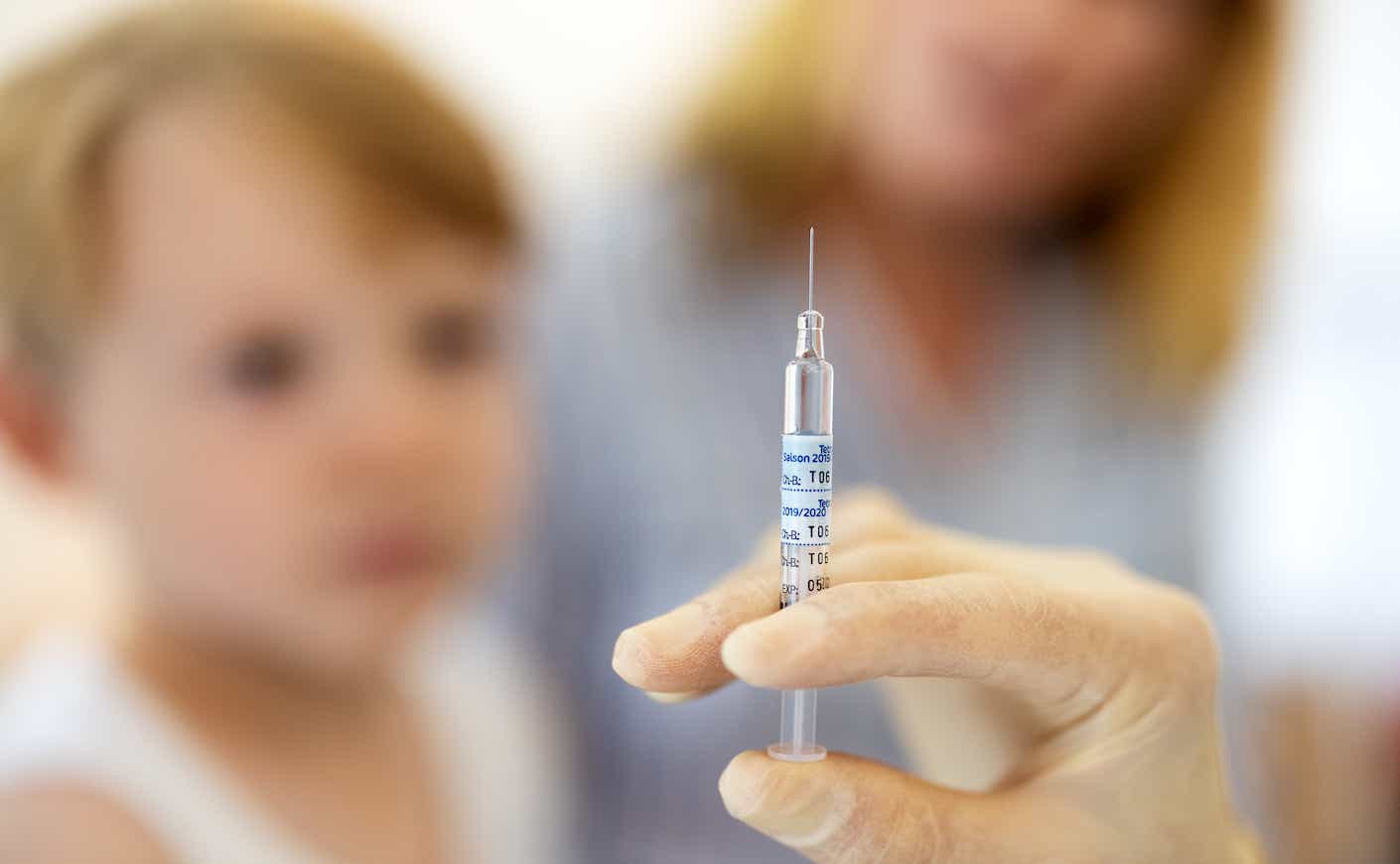 Close up of doctor preparing syringe for vaccine in clinic with a boy sitting at back. Hands of a pediatrician preparing flu injection.