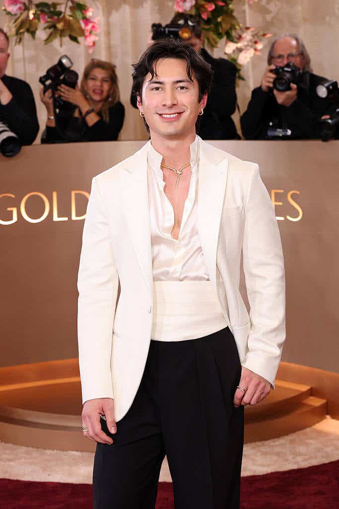 Hudson Williams wears a shiny white button-down with a white tuxedo jacket and white cummerbund and black pants.