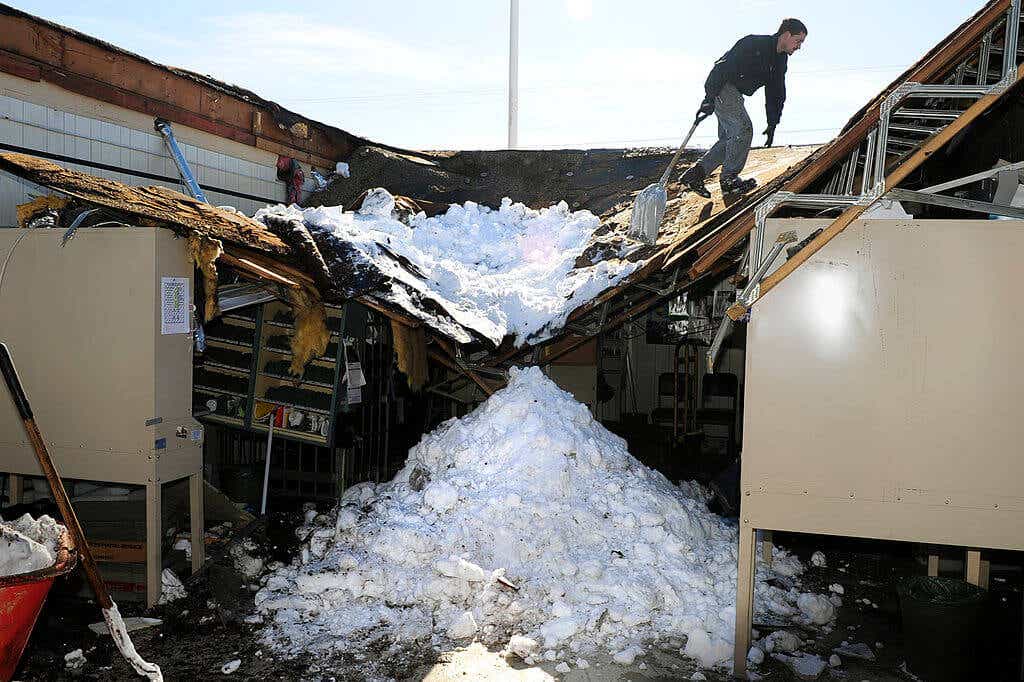 A worker with a shovel tending to a roof that has collapsed under snow.
