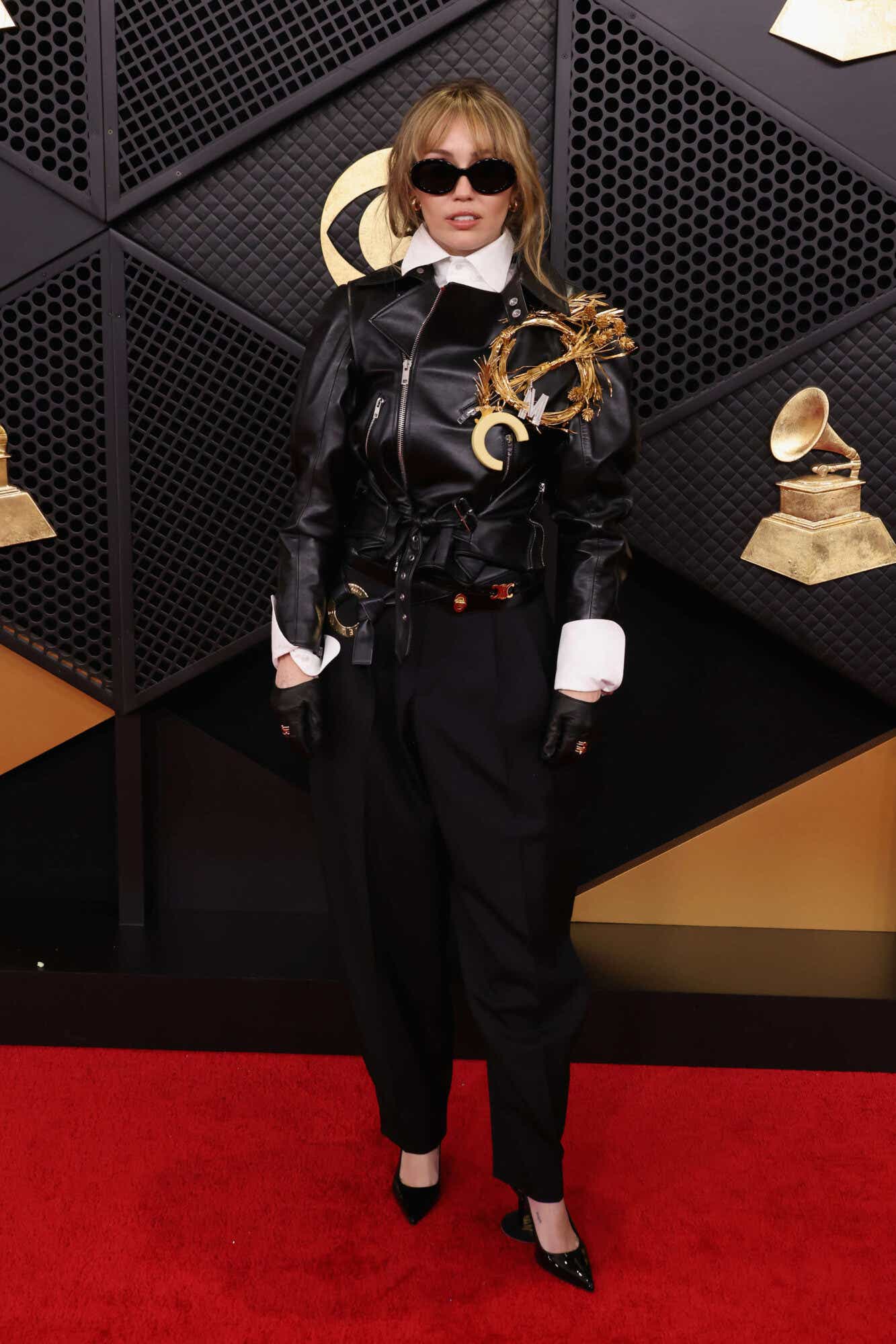 Miley Cyrus wears a leather jacket over a white button-down and a huge Mockingjay-esque brooch on the lapel