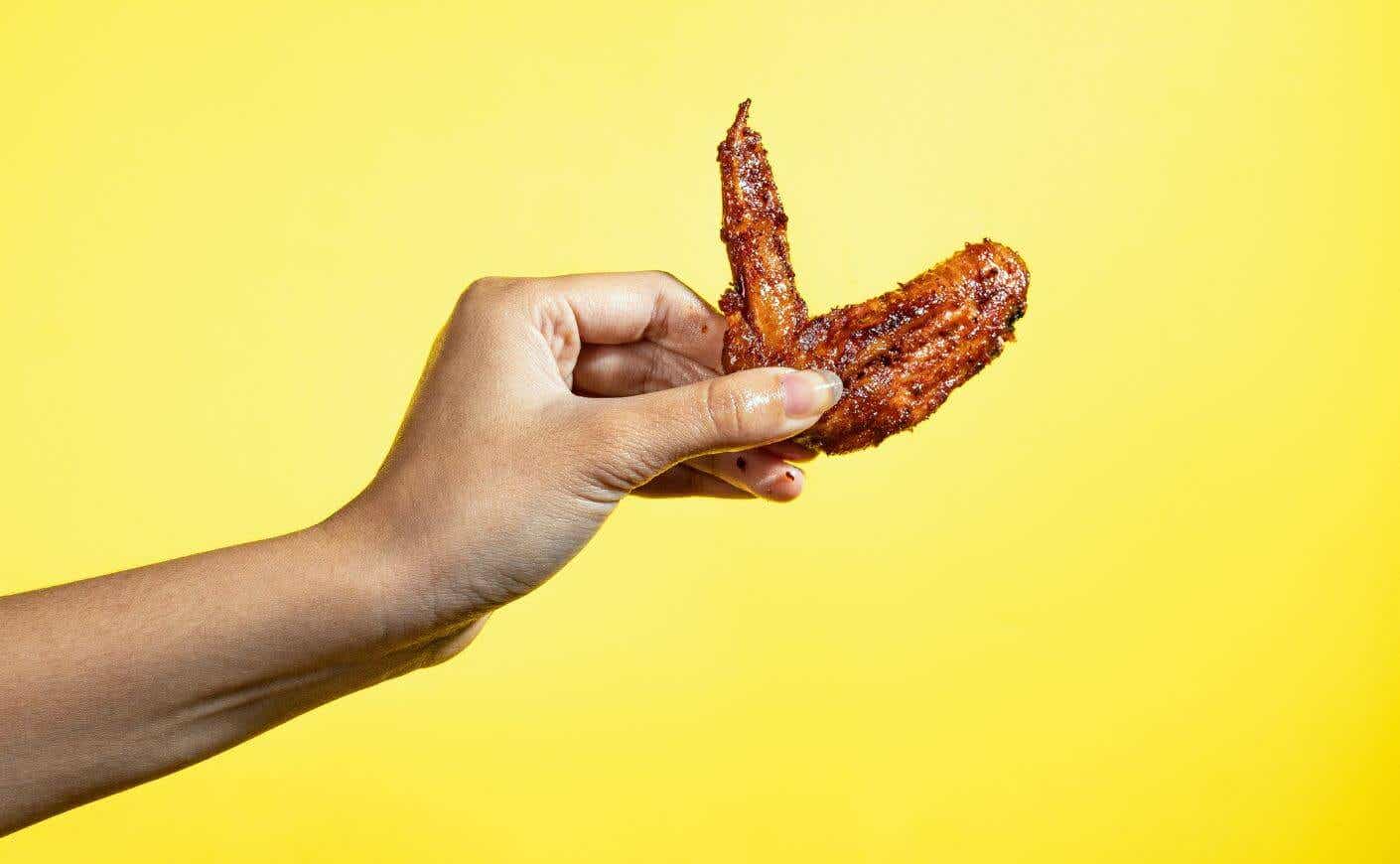 A hand holding up a Buffalo chicken wing against a yellow background.