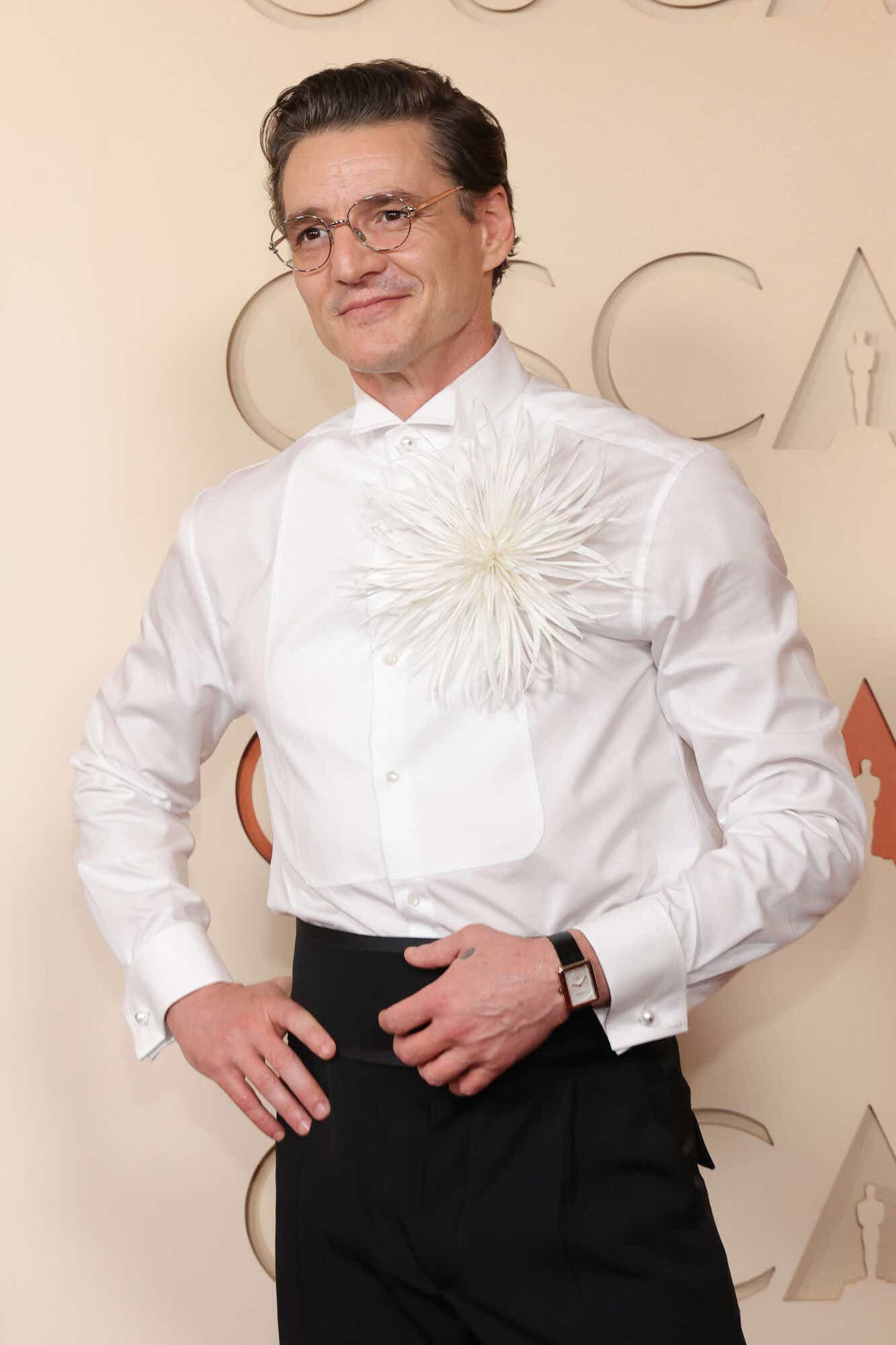 Pedro Pascal wears black tuxedo pants with a cummerbund and a white button-down with a big fluffy white flower on the breast.
