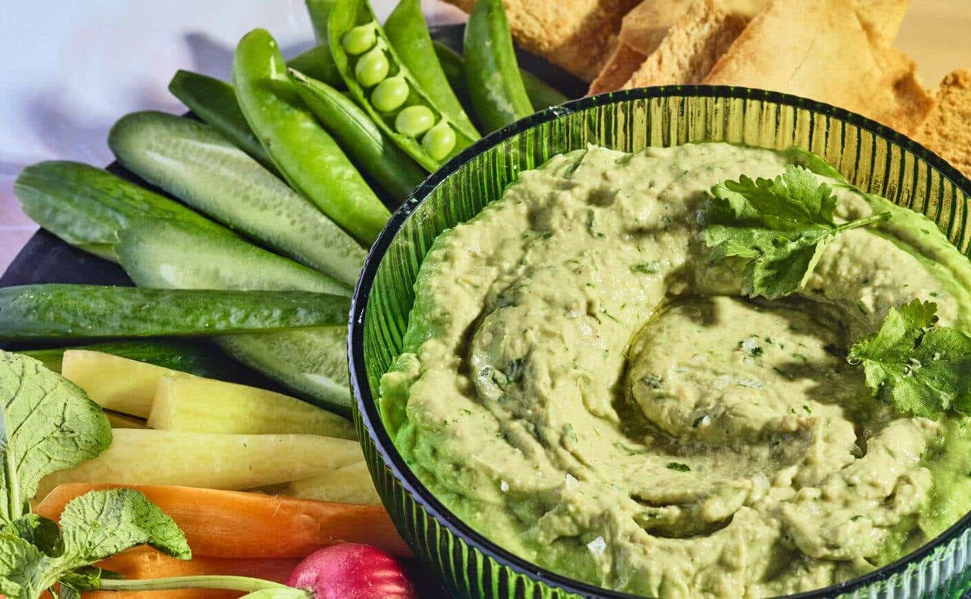Avocado hummus in a dish surrounded by vegetables and pita chips.