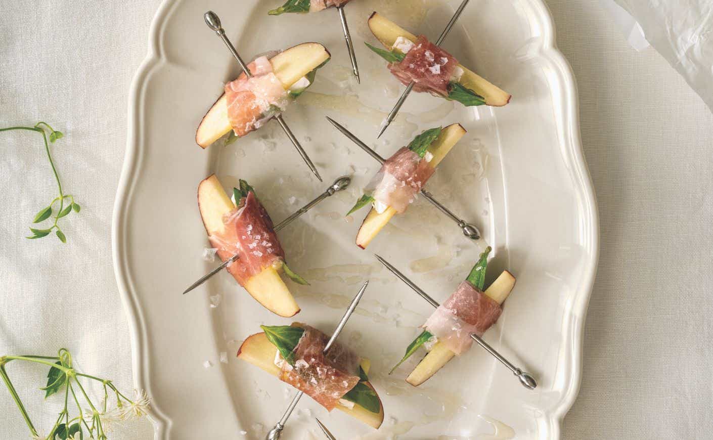 A platter of apple, brie, and prosciutto bites on metal toothpicks.
