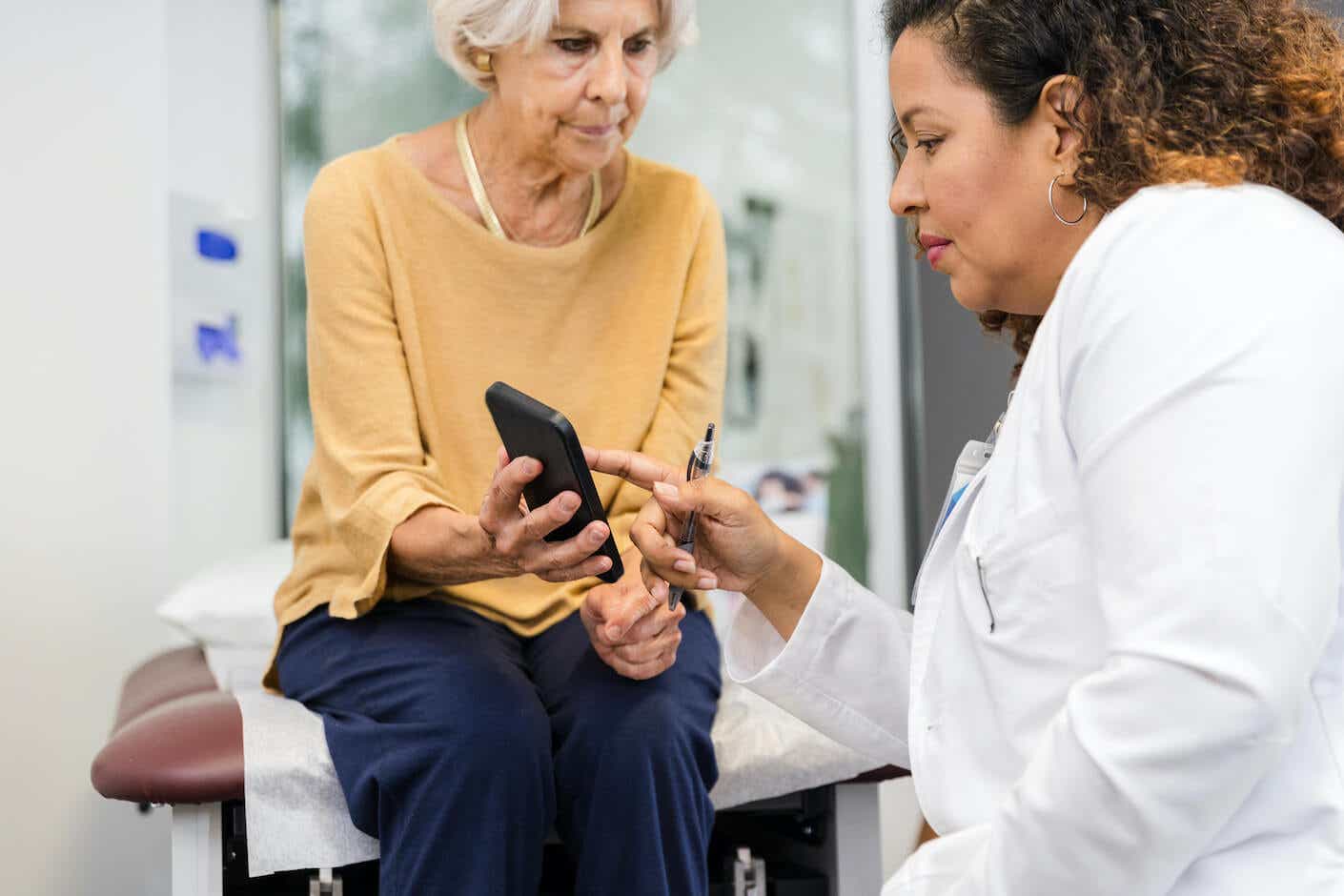 A senior adult woman uses her smartphone to ask her female doctor about the possibility of taking a medication she found advertised on the internet.