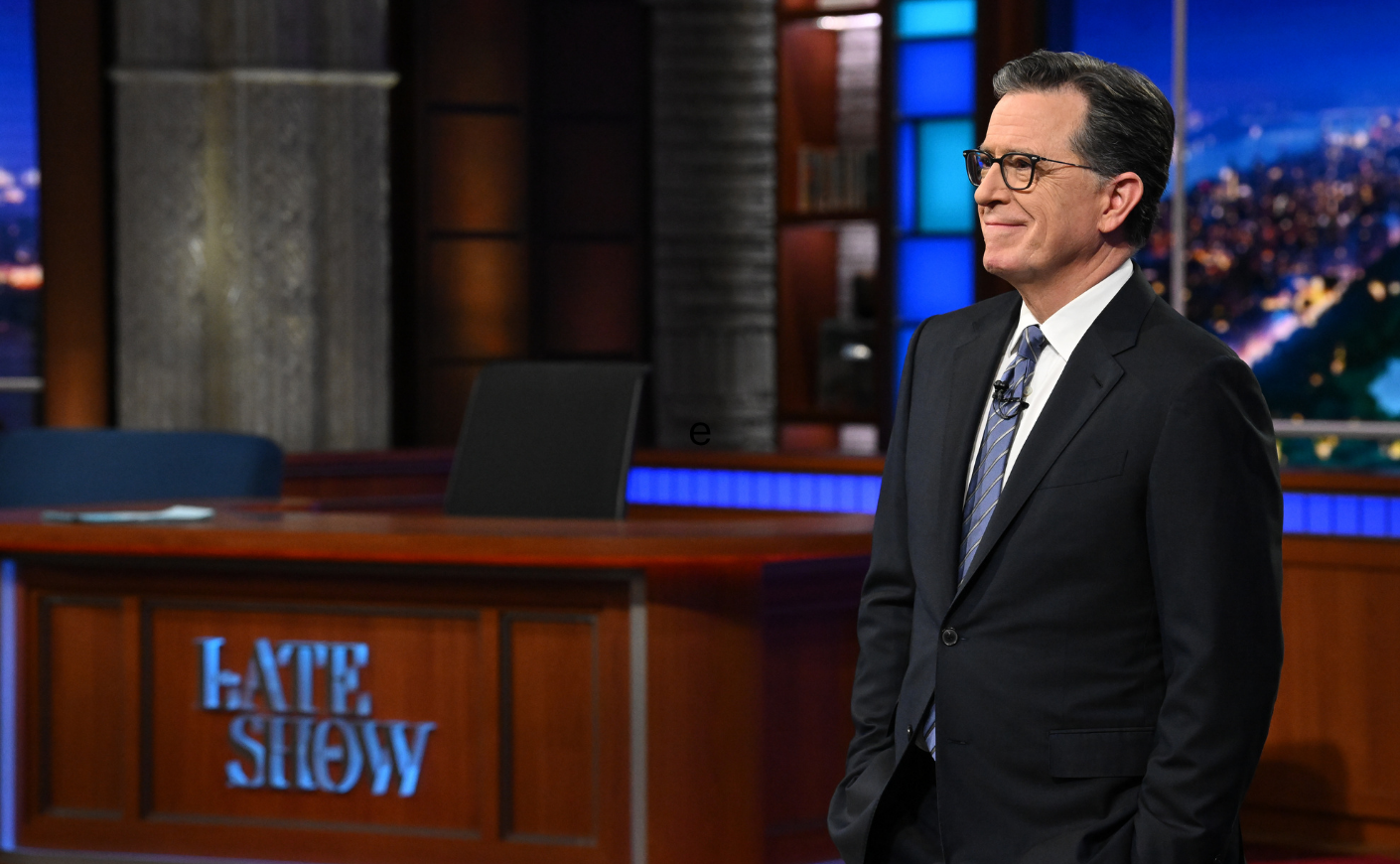Stephen Colbert wearing a suit and tie stands with his hands in his pockets in front of the Late Show desk, his hands in his pockets and a soft smile on his face.