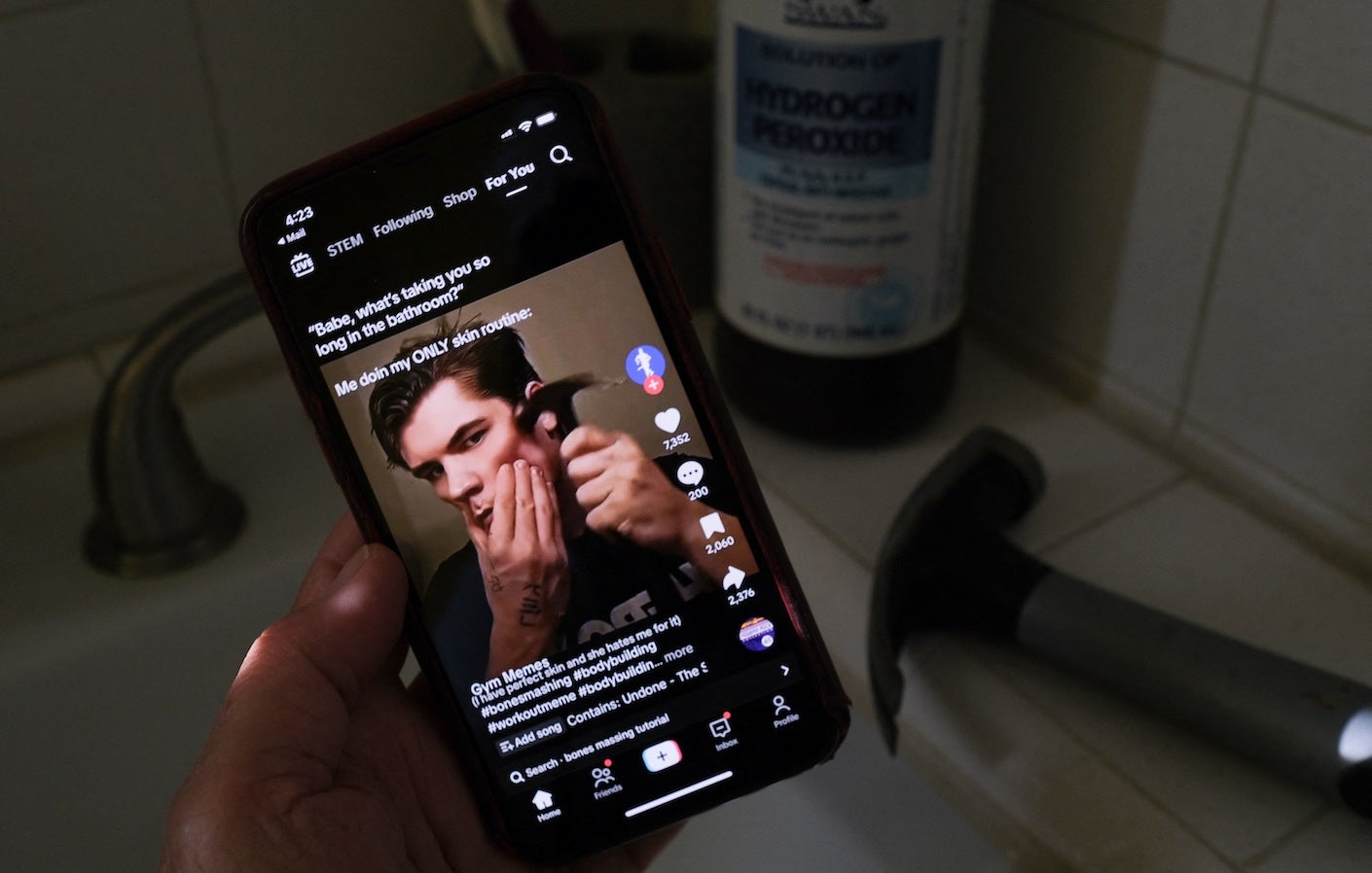 This photo shows a video of male TikTok influencer hammering his cheekbone, playing on a smartphone in a bathroom next to a bottle of hydrogen peroxide and a hammer