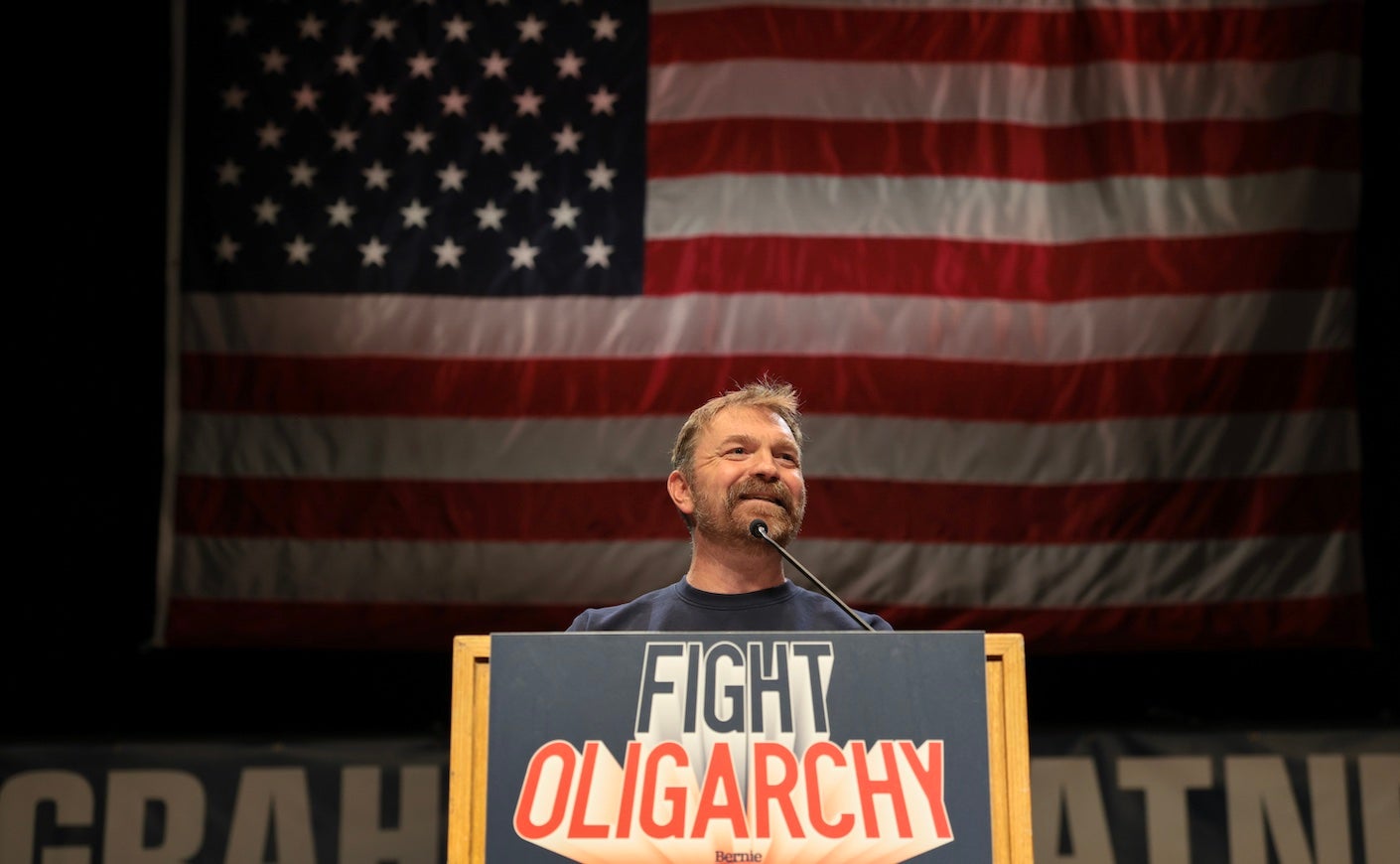 grahm platner speaking at a podium that says "fight oligarchy" with an american flag behind him