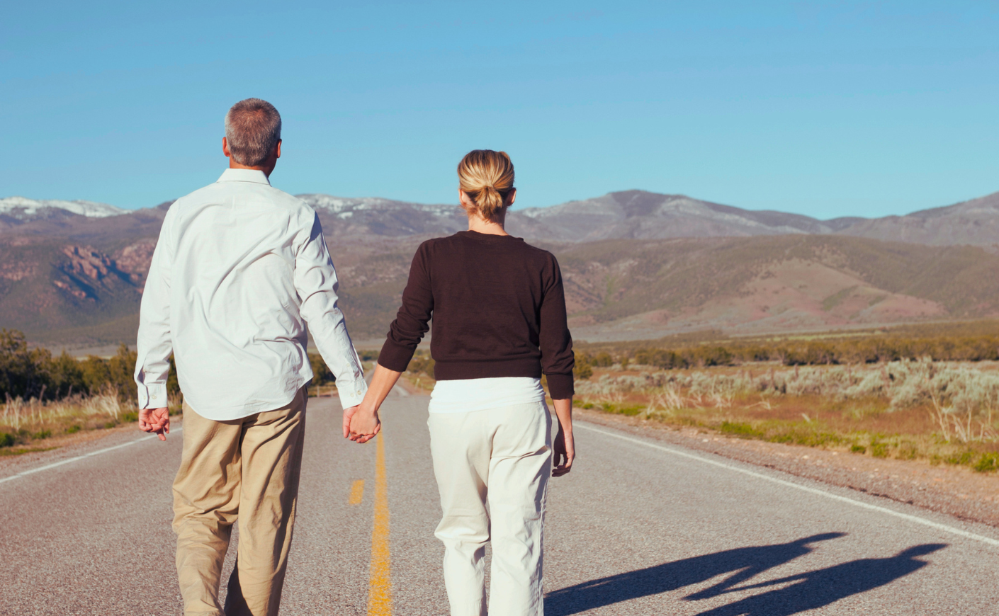 man and woman holding hands walking down a road toward mountains