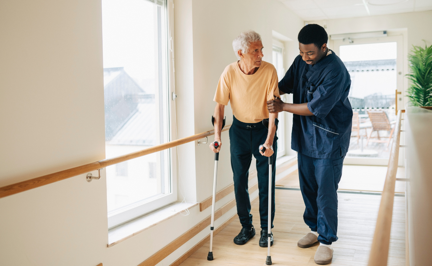 A Black male medical professional in navy scrubs helping an elderly white man walk up a ramp with crutches