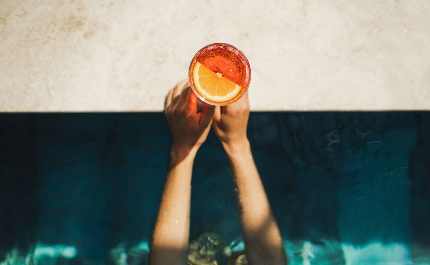 Two hands holding a drink on the edge of a pool.