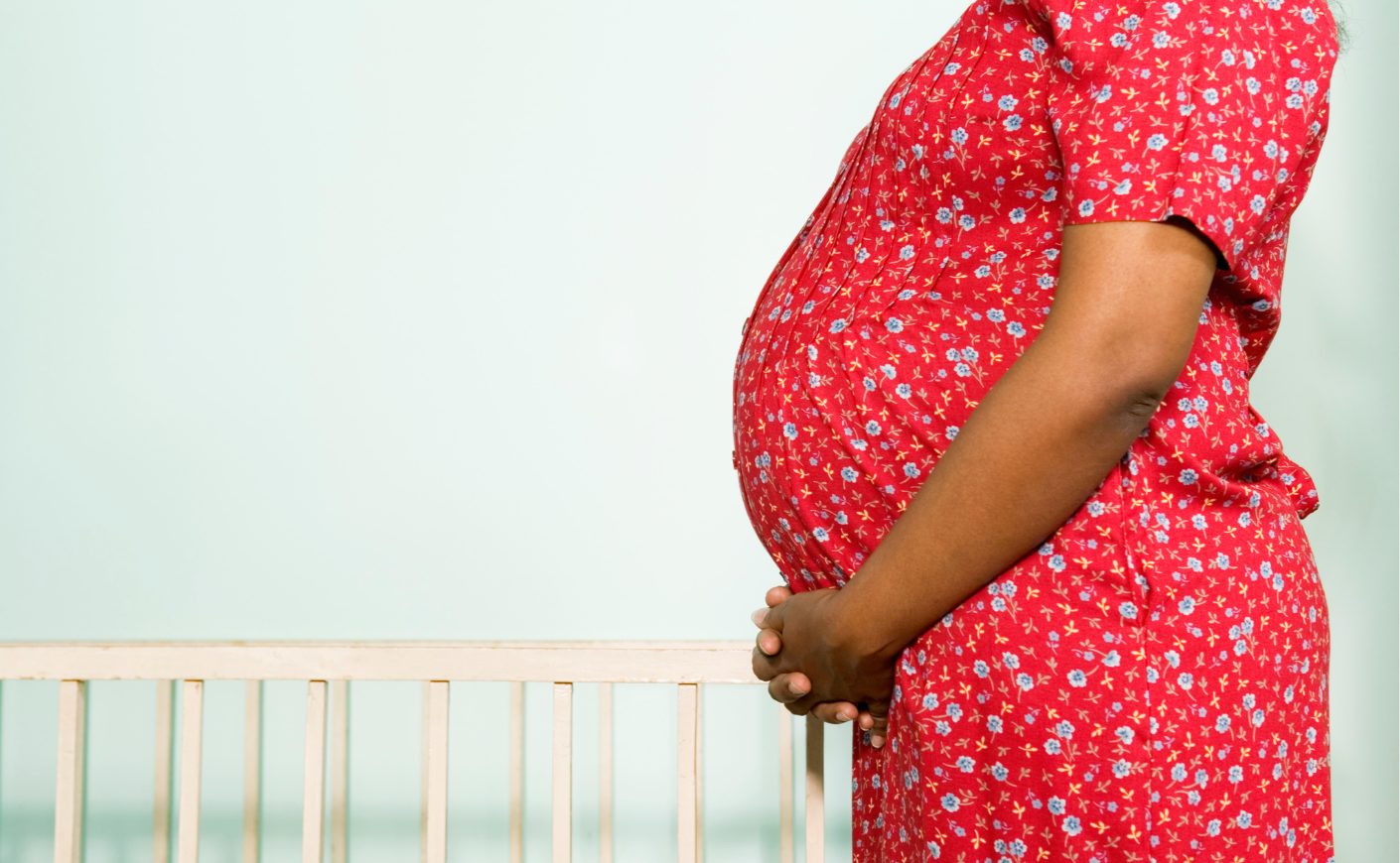 A black woman in a flowered red dress cradling her pregnant belly; a crib is visible in the background