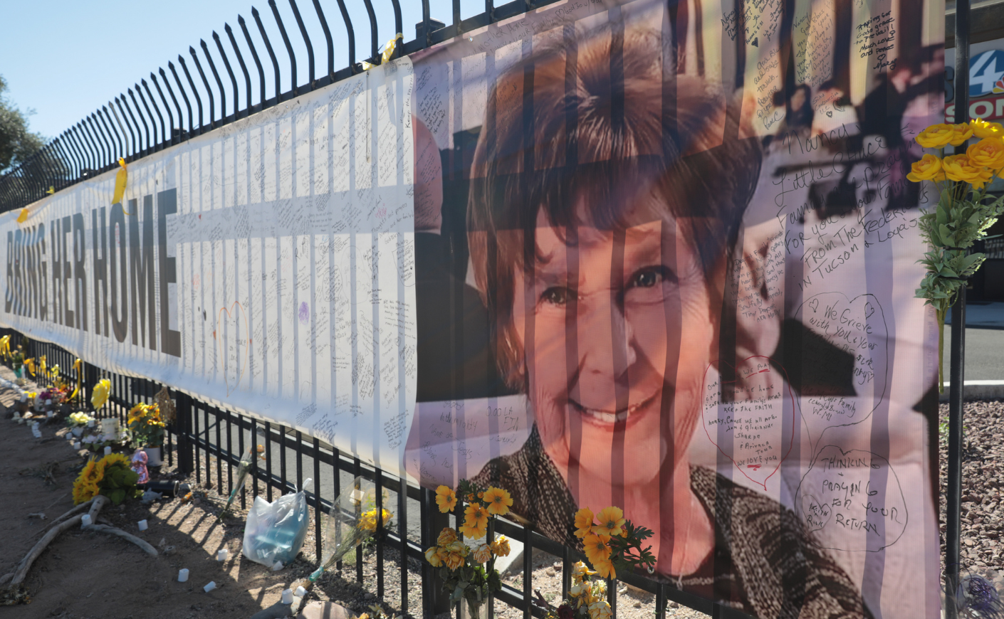 A banner stretched across a fence with a picture of Nancy Guthrie and the words BRING HER HOME