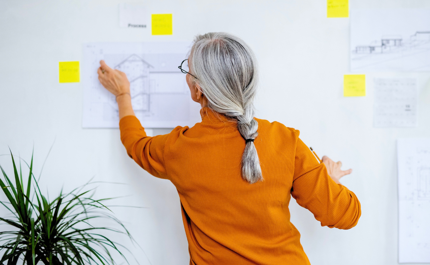 Woman in a orange sweater with gray hair pointing at renderings on a wall