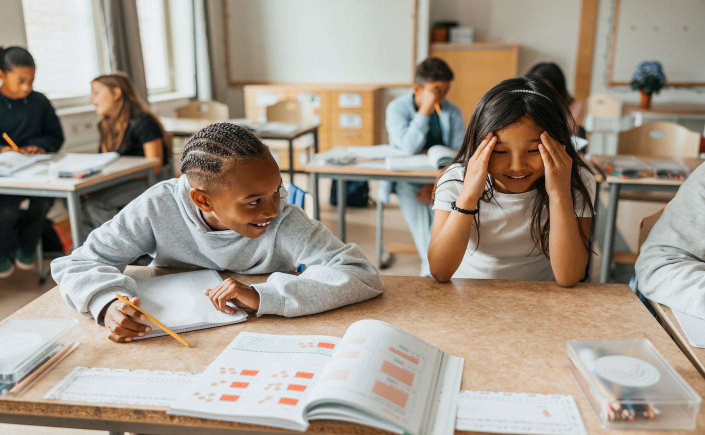 Smiling boy with braided hair sitting beside female friend touching temples while sitting at desk in classroom at school