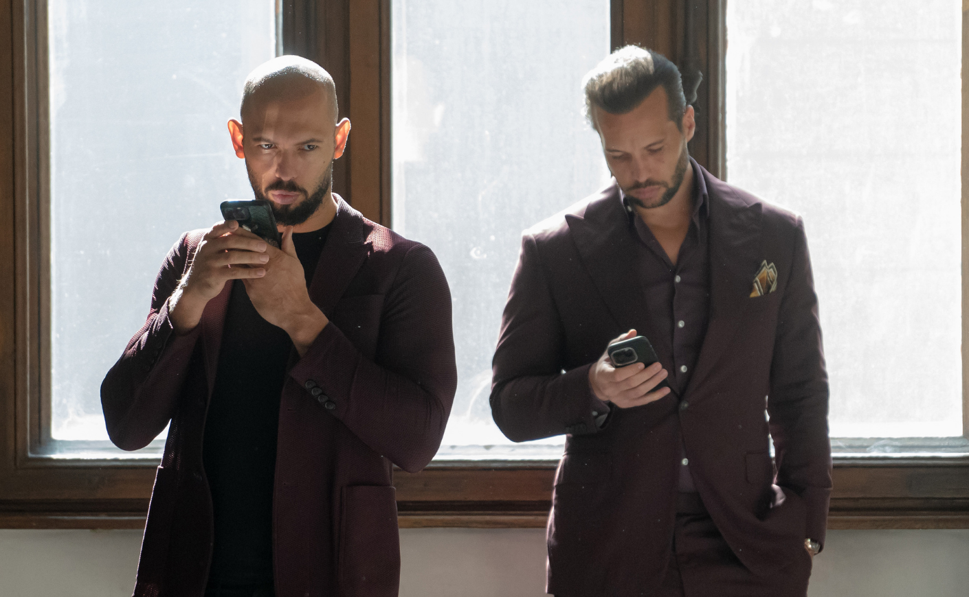 Andrew and Tristan tate leaning against a window, scrolling on their phones